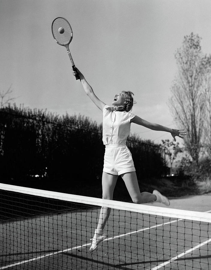 Black And White Photograph - 1950s Woman Jumping To Hit Tennis Ball by Vintage Images