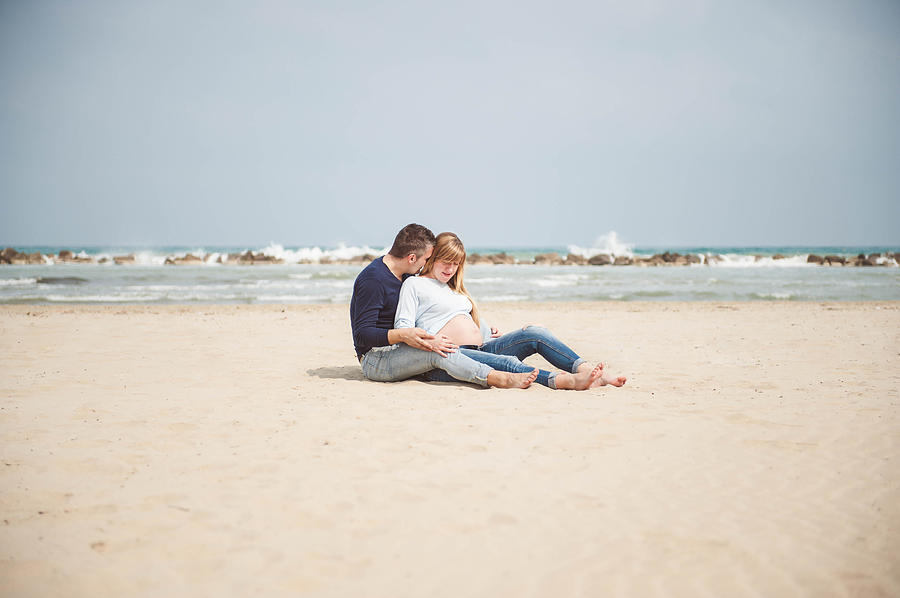 Pregnant couple sitting on the beach. Embrace. Casual clothes. #2 Photograph by © Samantha Carrirolo
