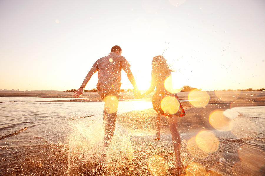 A happy couple runs through waves on sunlit beach Photograph by Vsurkov