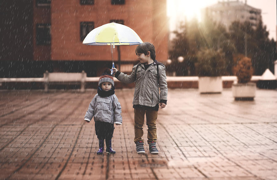 A little boy and his older brother protect themselves from the rain with an umbrella, in the city Photograph by Estersinhache fotografía