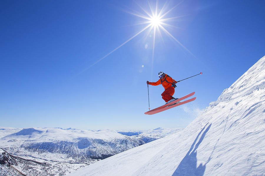 A male freerider in a red suit is jumping from a snow ridge. The sun is shining, the sky is blue.   Photograph by Menno Boermans