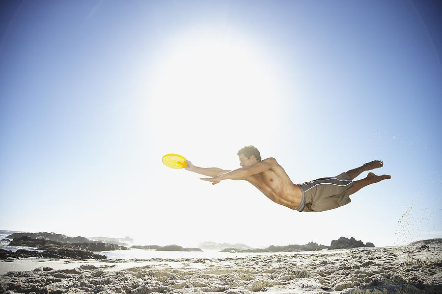 A man leaping through the air at the beach catching a Frisbee Photograph by OJO Images