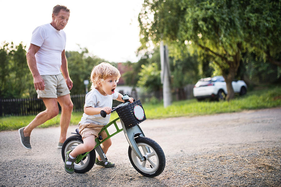 A small toddler boy with his grandfather riding a balance bike outdoors. Photograph by Halfpoint Images