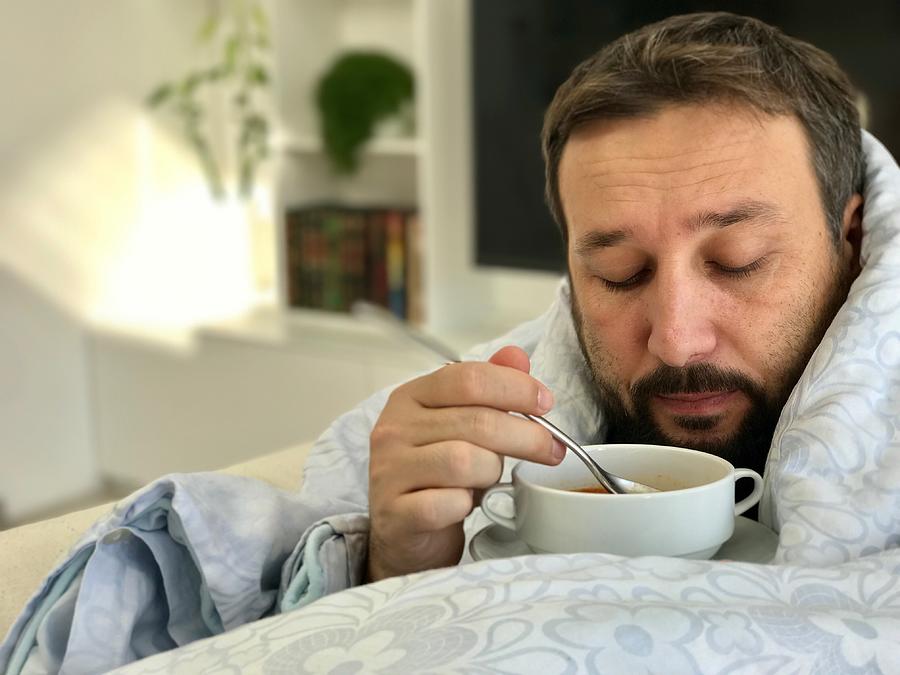 Adult man lying sick on sofa in living room eating soup with no taste Photograph by Jasmin Merdan