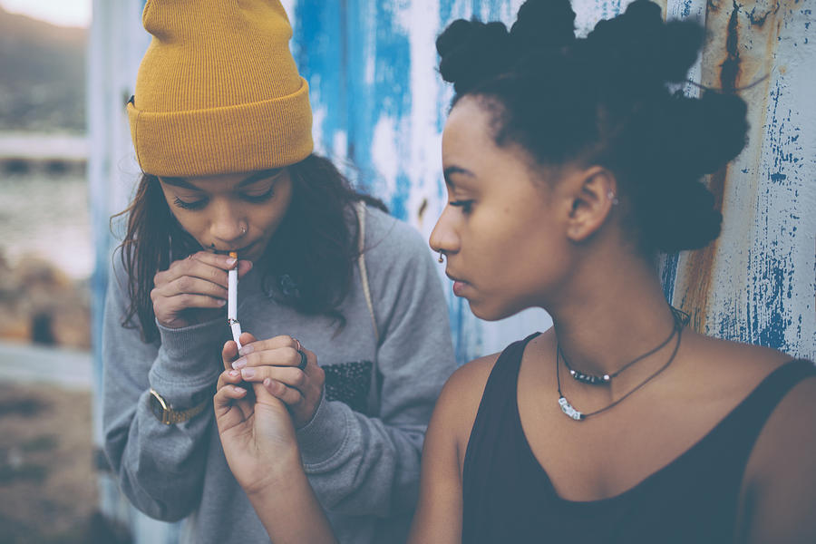 Afro teen lighting a cigarette for her grungy girl friend Photograph by Wundervisuals