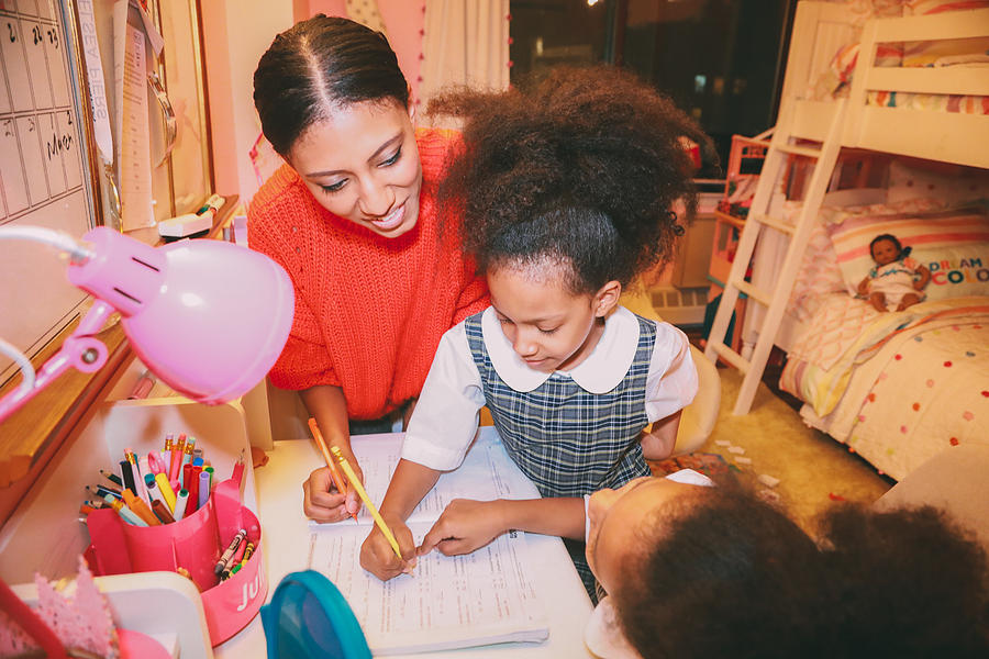Afrolatina mom and twin daughters doing homework at home. Daughters are in their school uniform. Background is a pink bedroom. Photograph by Evelyn Martinez