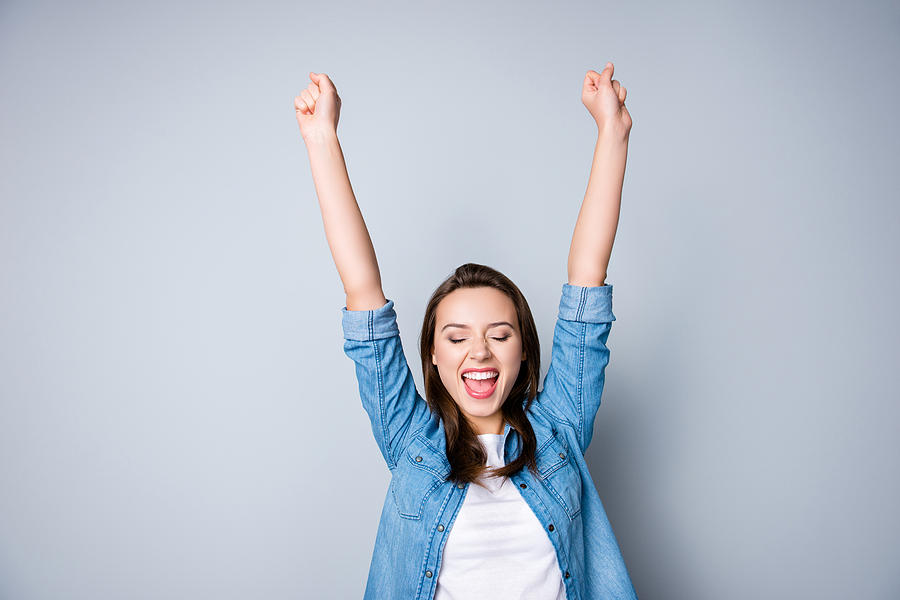 Amazed brunette young business woman in casual shirt is gesturing victory with her raised hands, she is shocked, extremely happy, with closed eyes, beaming smile, open mouth  on  grey background Photograph by Deagreez