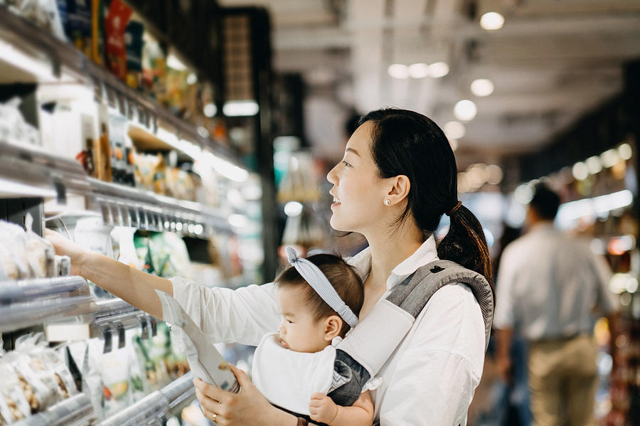 Beautiful Asian woman carrying cute baby girl shopping for organic green marketing products in grocery store Photograph by D3sign
