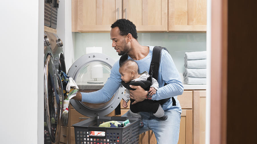 Black father with son in baby carrier doing laundry Photograph by Ariel Skelley