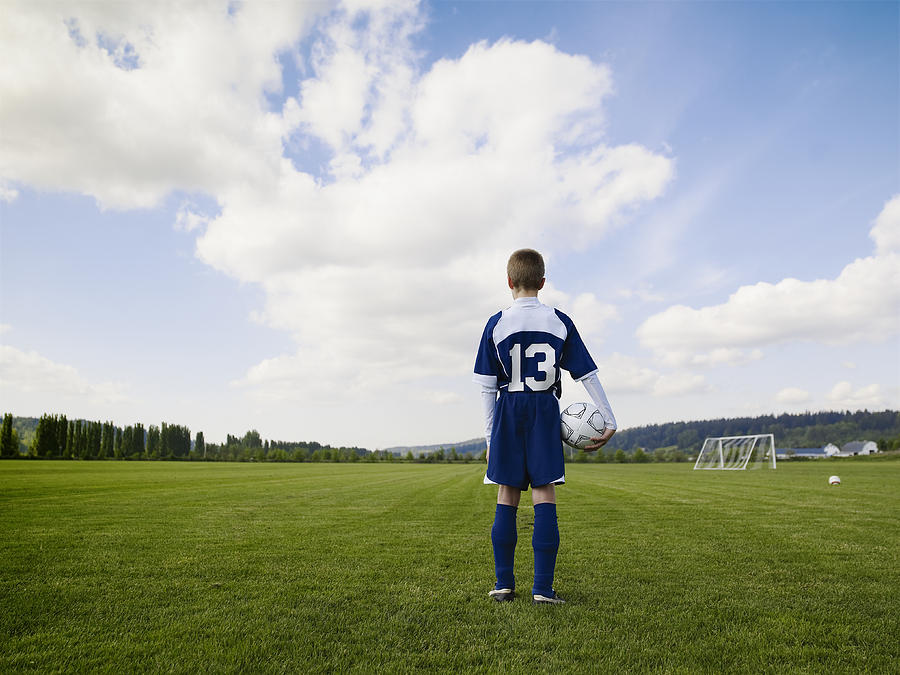 Boy (12-13) standing, holding football, on pitch, rear view Photograph by Thomas Barwick