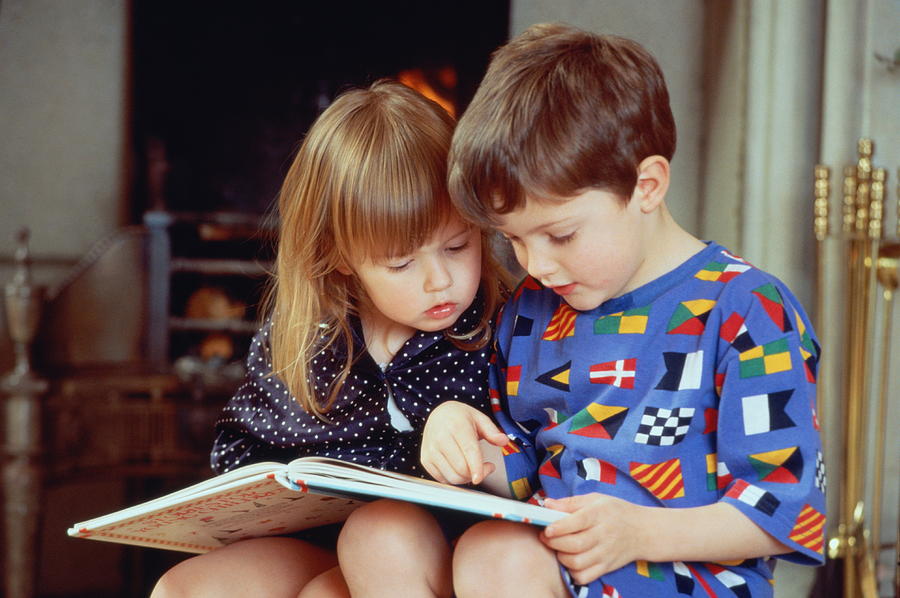 Boy(4-5)and girl(2-3)sat by fire in pyjamas reading book together Photograph by Bob Thomas