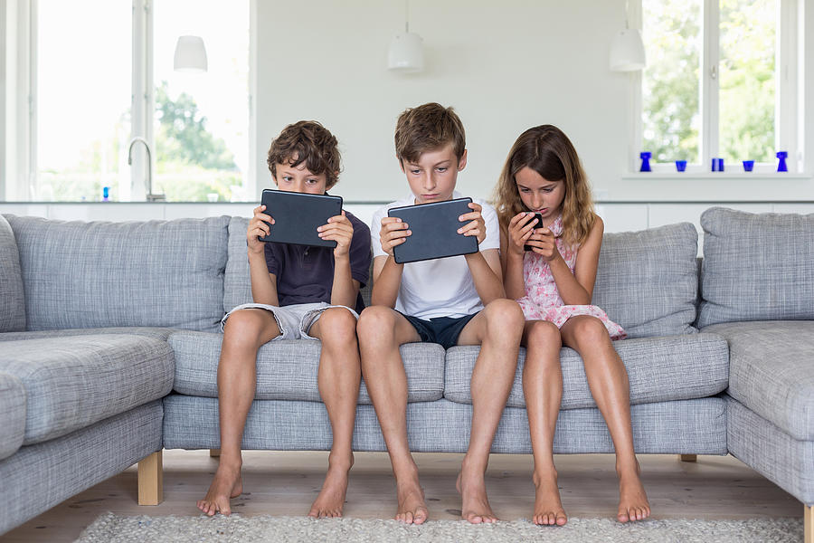 Brothers and sister on sofa with digital tablets and mobile Photograph by Richard Lewisohn