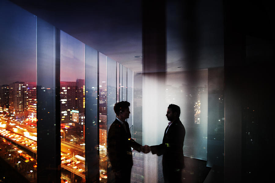Businessmen shaking hands in office at night with city view Photograph by FangXiaNuo