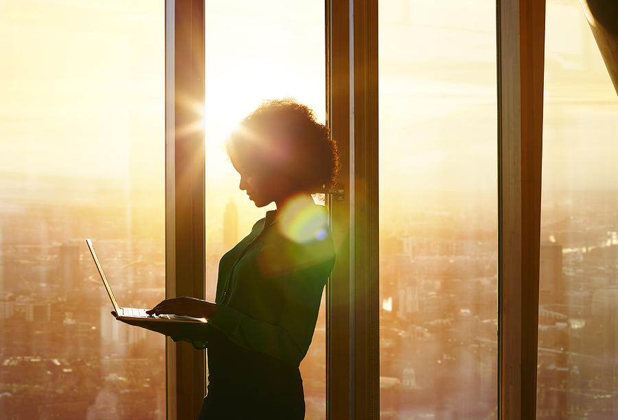Businesswoman on laptop at window in morning sun Photograph by Ezra Bailey