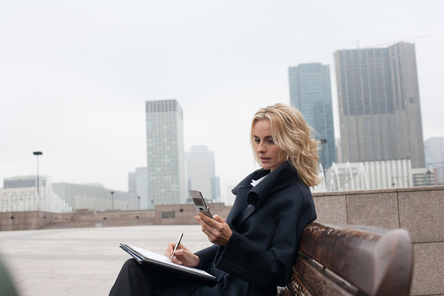 Businesswoman outdoors writing in notebook with celluar phone Photograph by Sam Edwards