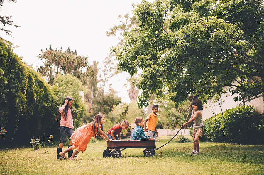 Children pushing and pulling friends in red wagon in park Photograph by Wundervisuals