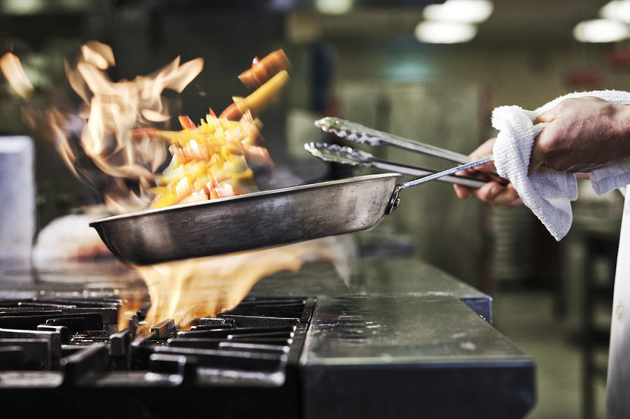 Close-up of chefs hands holding a saute pan to cook food, flambeing contents. Flames rising from the pan. Photograph by Mint Images