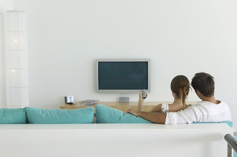 Couple Sits on Sofa, Woman Pointing a Remote Control at Plasma Television Photograph by Flying Colours Ltd
