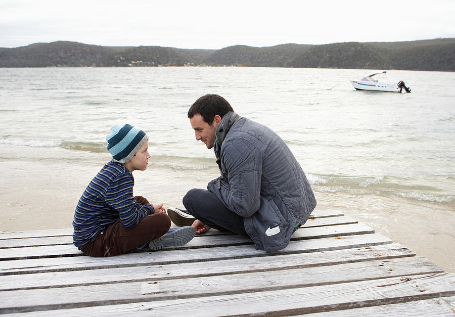 Father and son (10-11) sitting at end of dock at edge of lake, talking, side view Photograph by Marc Debnam