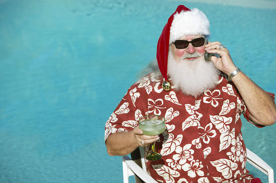 Father Christmas Wearing a Hawaiian Shirt Sitting by a Swimming Pool Holding a Mobile Phone and a Cocktail Glass Photograph by Digital Vision.