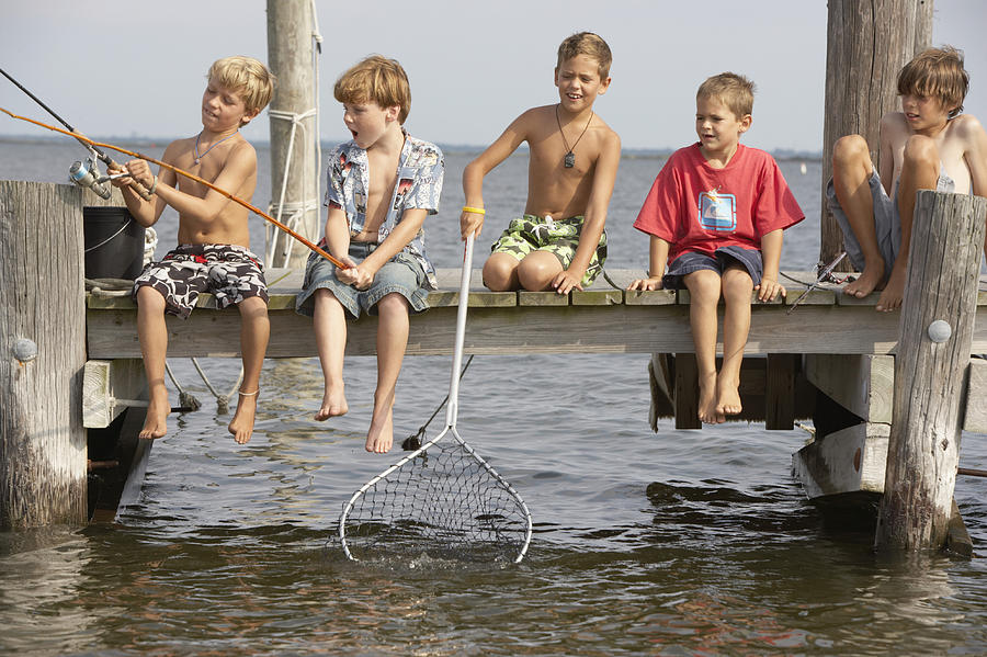 Five boys (5-13) sitting on jetty fishing, one holding net in water Photograph by Christopher Robbins
