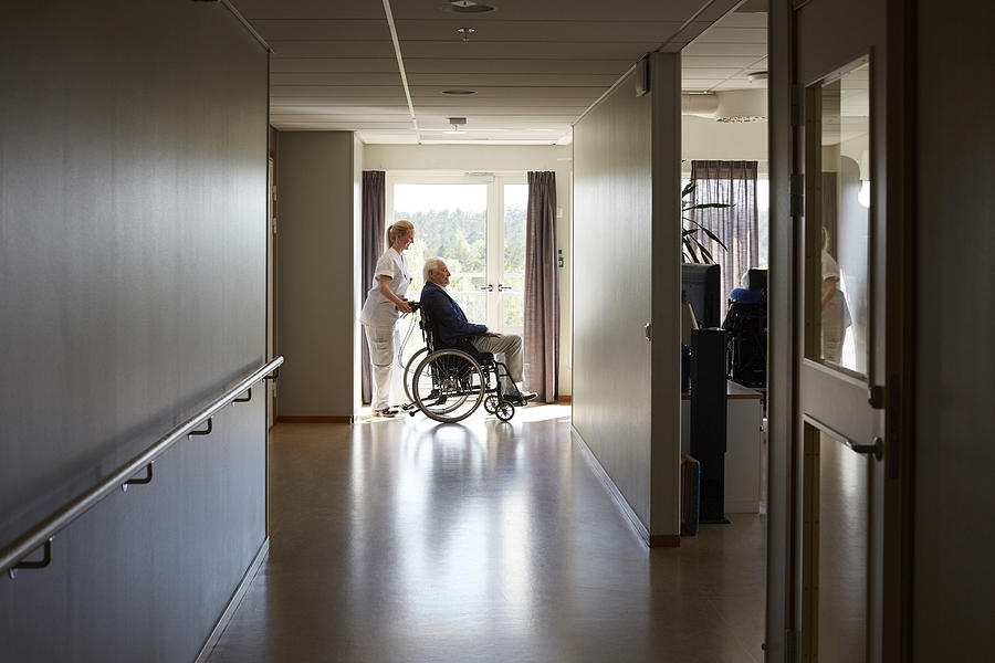 Full length side view of female nurse pushing senior man on wheelchair at hospital corridor Photograph by Maskot