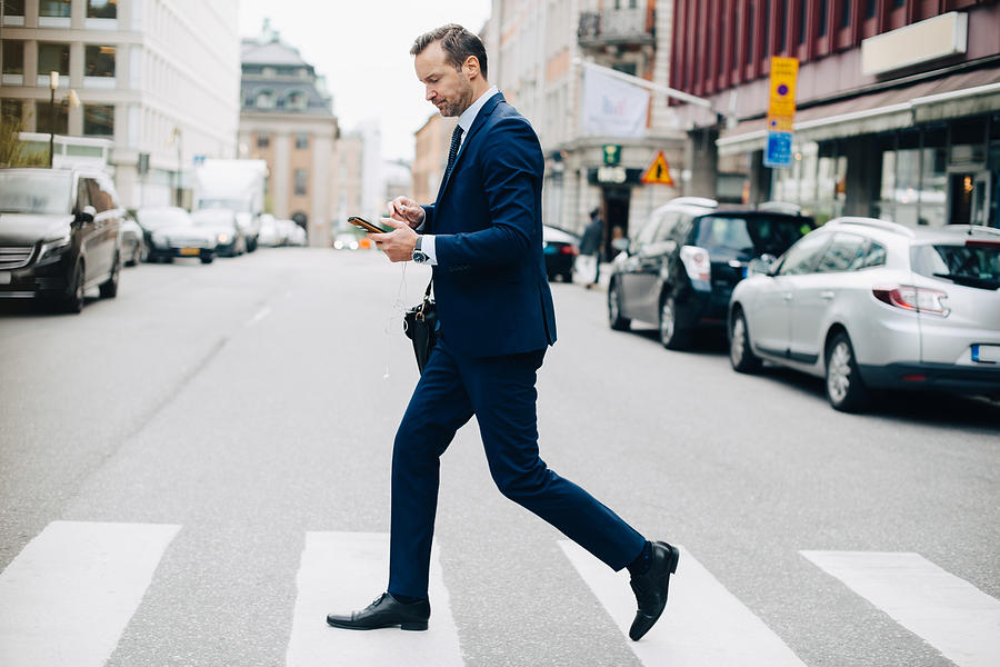 Full length side view of mature businessman crossing street while using smart phone in city Photograph by Maskot