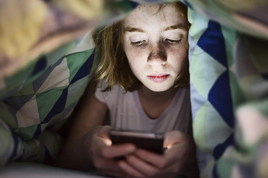 Girl lying under blanket in bed looking at her smartphone Photograph by Westend61