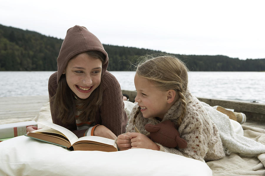 Girls (6-13 years) lying on jetty beside lake, reading book together and smiling Photograph by Noel Hendrickson