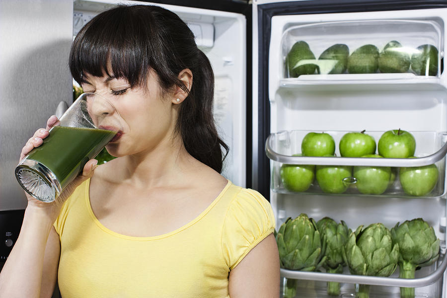 Grimacing mixed race woman drinking healthy drink near refrigerator Photograph by Jill Giardino