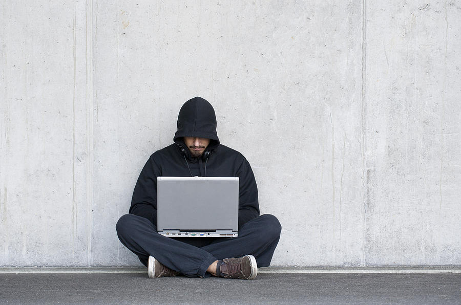 Hacker with laptop sitting in front of concrete wall Photograph by Westend61