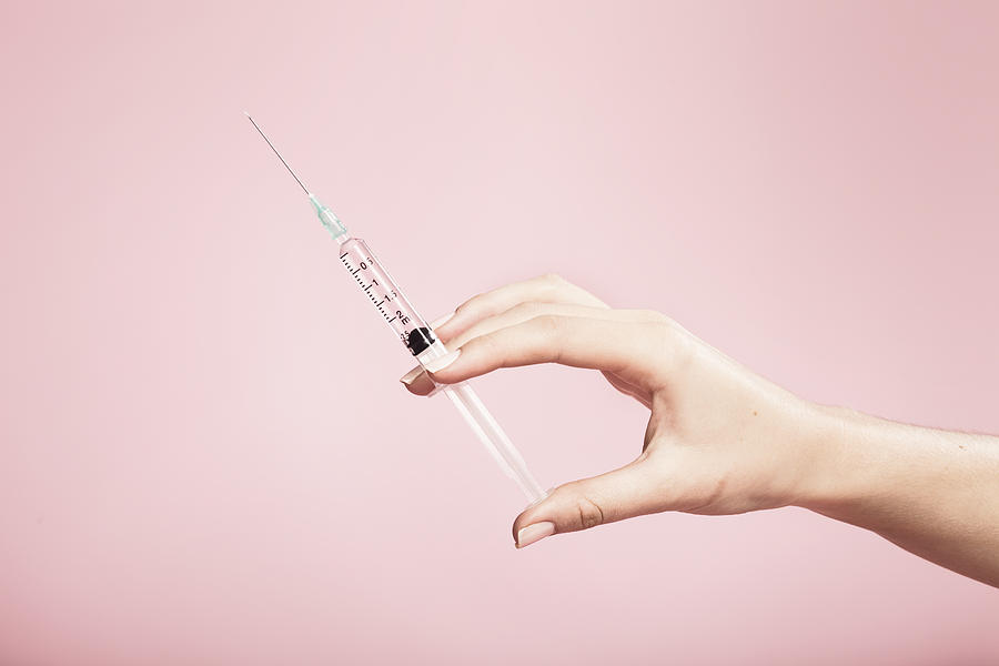 Hand holding syringe in plain pink background Photograph by Paper Boat Creative