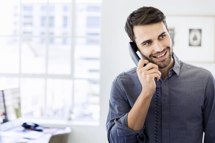 Happy businessman using landline phone in office Photograph by Portra