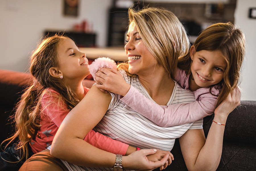 Happy mother enjoying in a hug by her two little daughters at home. Photograph by Skynesher