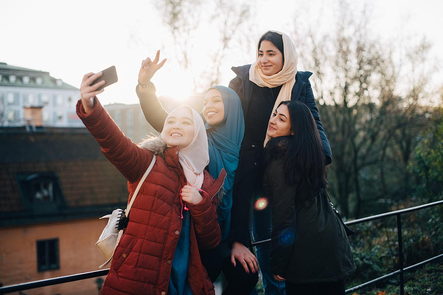Happy young Muslim woman taking selfie with friends by railing in city Photograph by Maskot