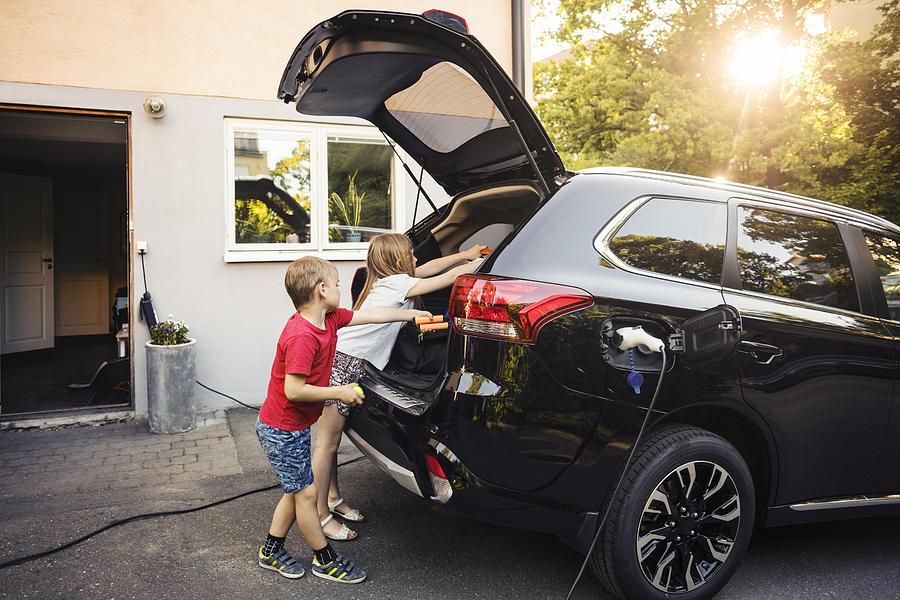 Kids loading black electric car trunk against house in back yard Photograph by Maskot