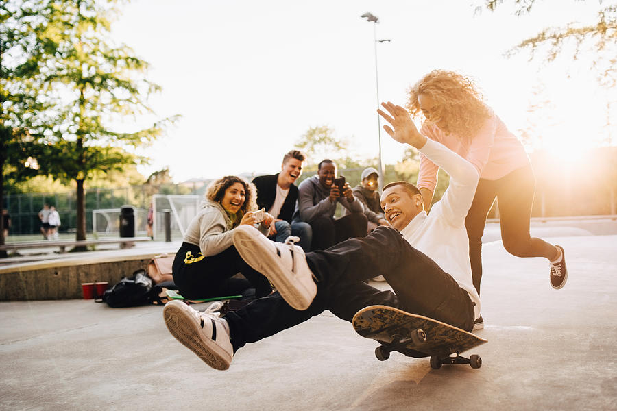 Laughing friends photographing man falling from skateboard while woman pushing him at park Photograph by Maskot