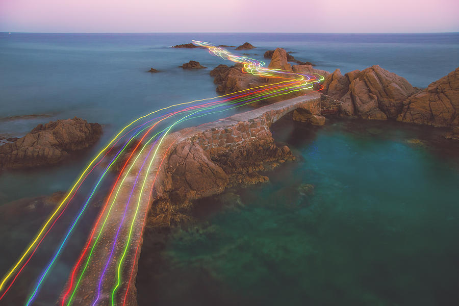 Long exposure of a colorful rainbow of light traces in a beautiful curved path between the rocks over the Mediterranean Sea in the Costa Brava shoreline on sunset. Photograph by Artur Debat