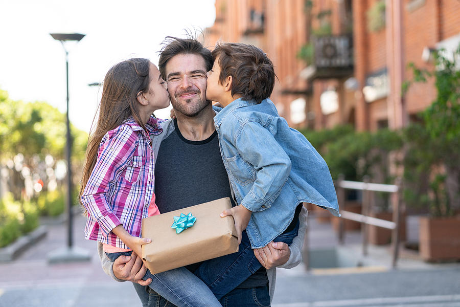 Loving son and daughter kissing daddy on the cheek while he carries them both with a present for fathers day Photograph by Hispanolistic