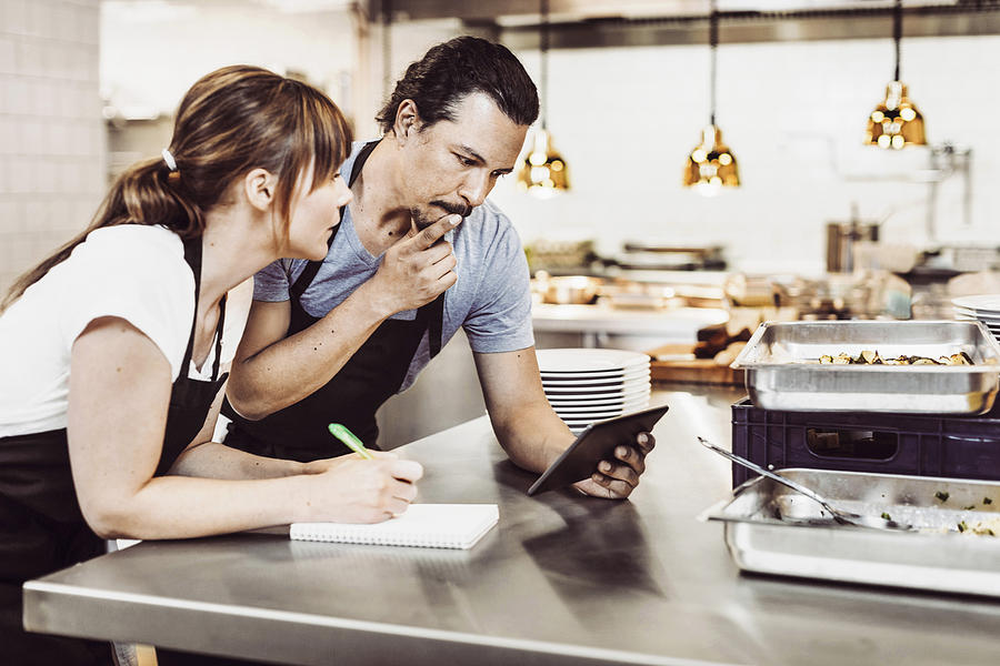 Male and female chefs using digital tablet while writing recipe at commercial kitchen counter Photograph by Maskot