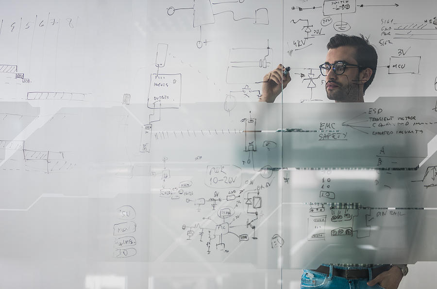 Male engineer working on new ideas and writing diagram on glass wall. Photograph by Skynesher
