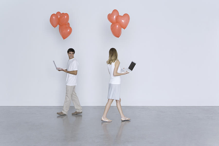 Man and woman walking past each other, both carrying laptop computers and heart balloons Photograph by PhotoAlto/Ale Ventura