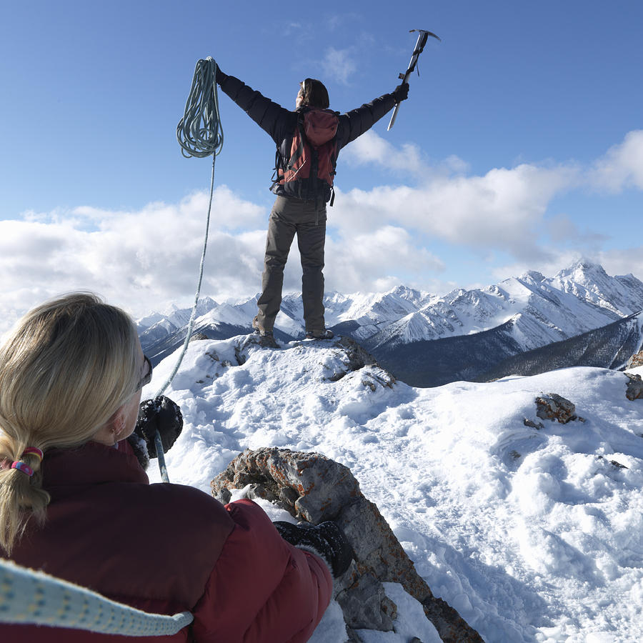 Man celebrating on mountain peak holding climbing equipments, woman in background, rear view Photograph by Ascent/PKS Media Inc.