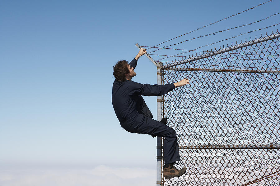 Man climbing chain link fence with barbed wire on top Photograph by Ryan McVay