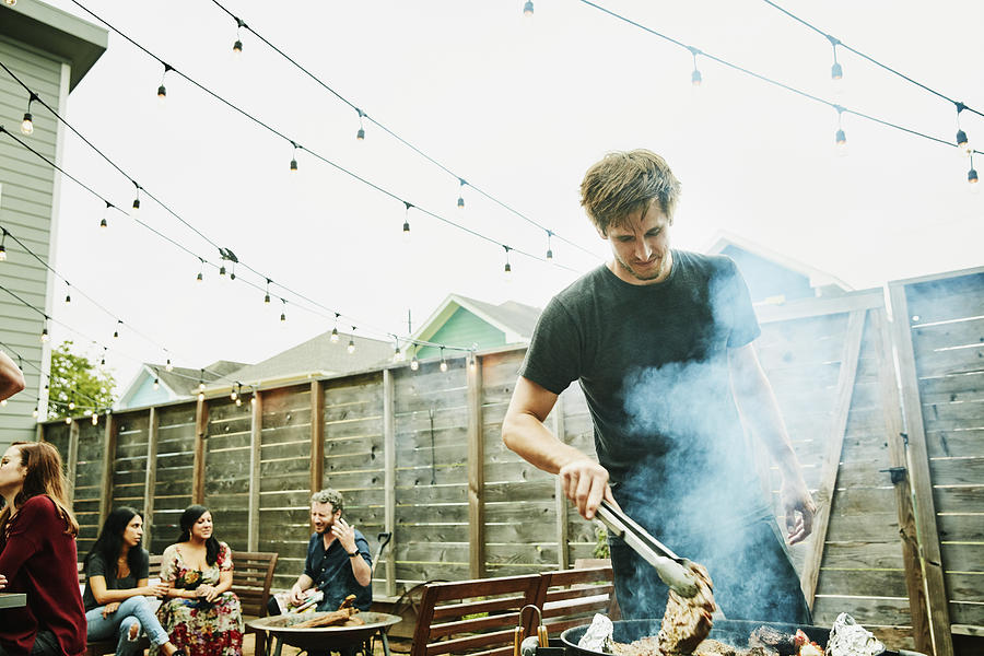 Man grilling steaks for friends at backyard barbecue during party Photograph by Thomas Barwick