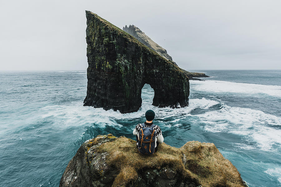 Man in knitted sweater looking at view of Drangarnir arch in Faroe Islands Photograph by Oleh_Slobodeniuk