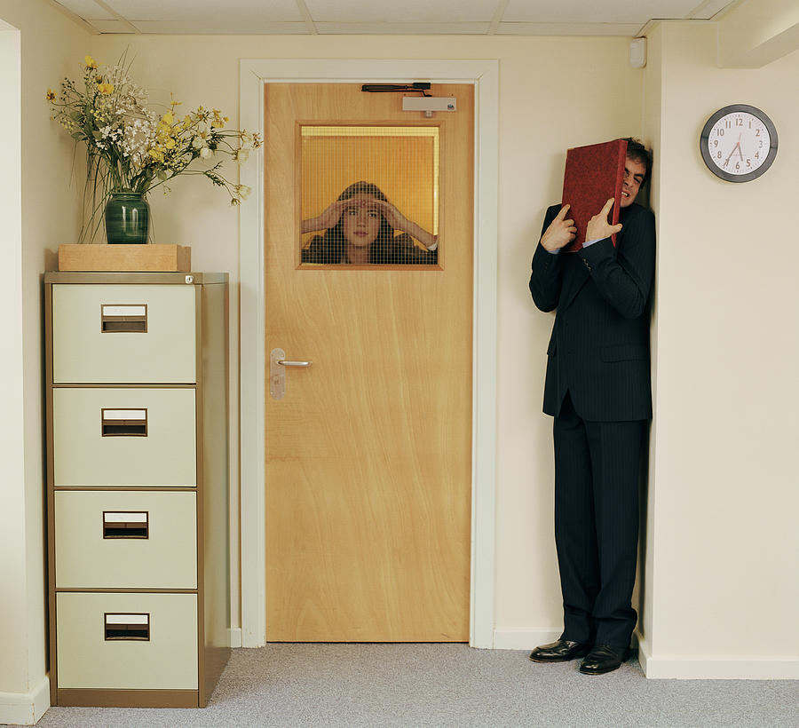 Man in office hiding from woman peering through window in door Photograph by Colin Hawkins