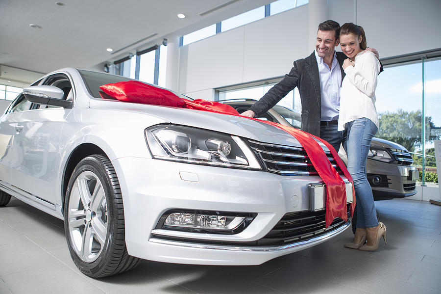 Man looking at new car with red bow with girlfriend in car dealership Photograph by Zero Creatives