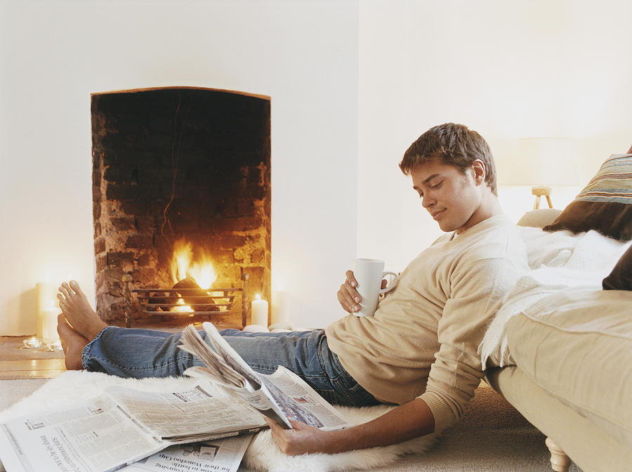 Man Sits on Rug by Fireplace Holding a Cup and Reading a Newspaper Photograph by Digital Vision.