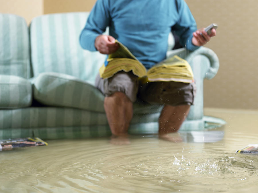 Man sitting in flooded living room using phone, low section Photograph by Michael Blann